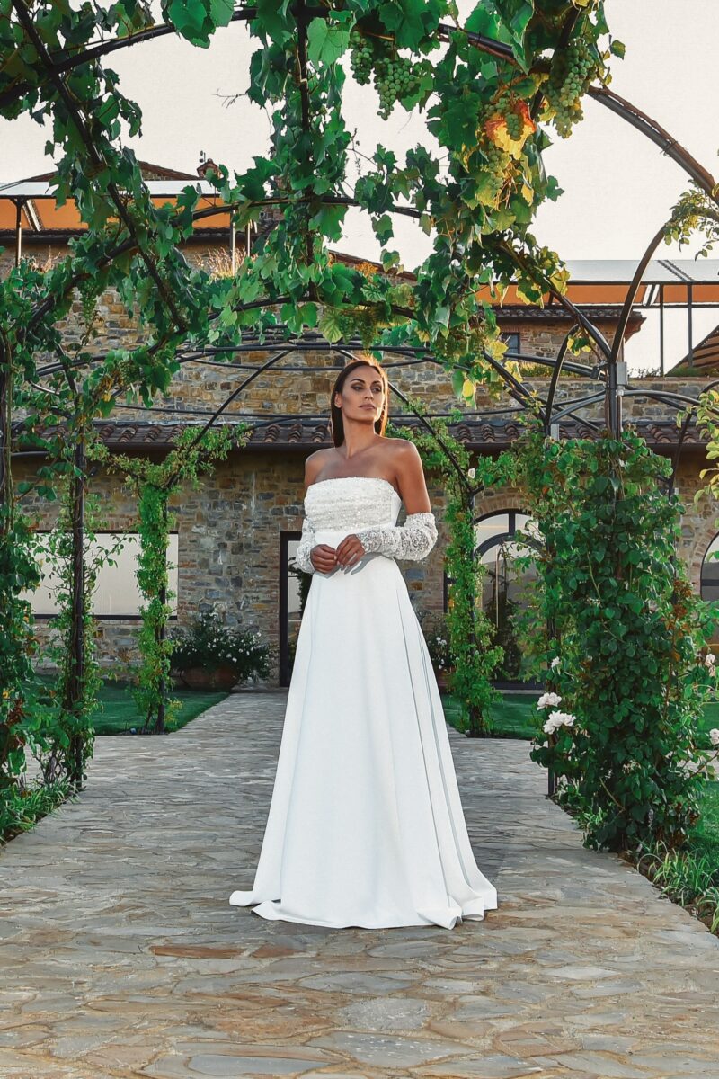 ORSOYA Menyasszonyi Ruha: Pánt nélküli minimal stílusú esküvői ruha, levehető félig áttetsző ujjrésszel, flitterrel, gyönggyel díszített dekoltázsrész.